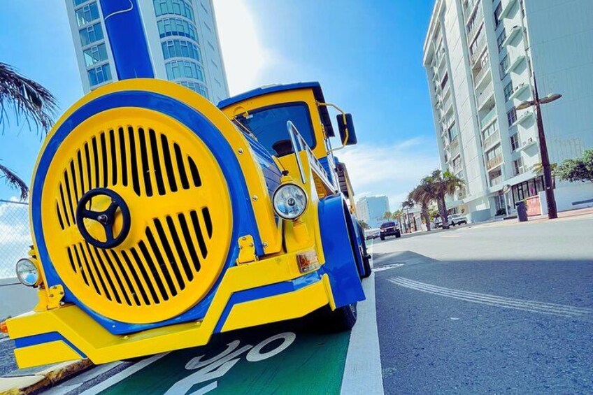 Een gele en blauwe tram rijdt over een stadsstraat, met gebouwen en palmbomen op de achtergrond. 2
