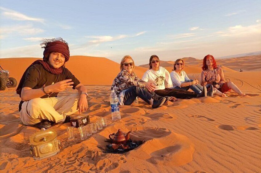 A group of people sitting on a sand dune with a tent and a small fire pit in the foreground. 2
