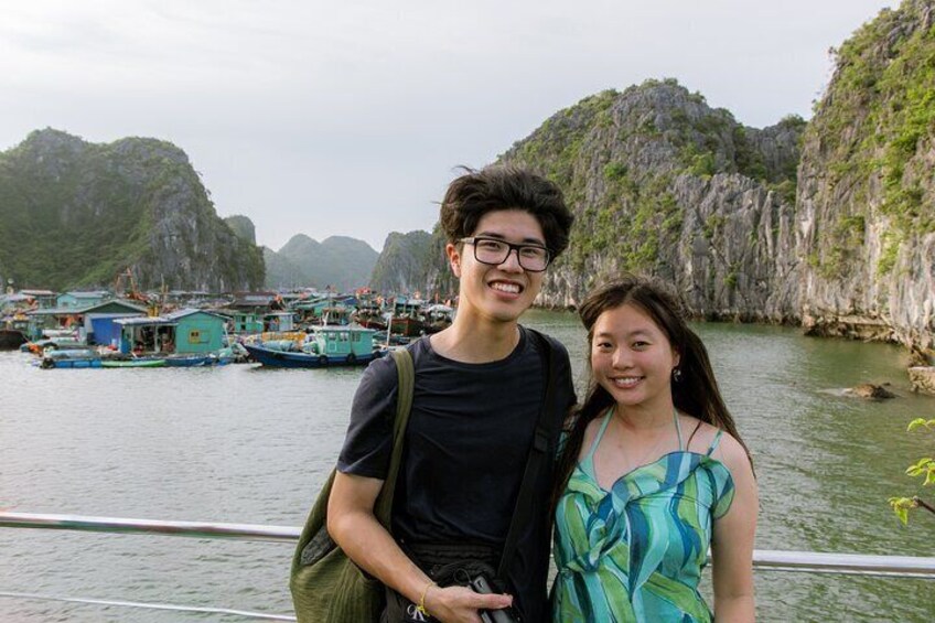 Two people standing by a railing with a view of boats and rocky cliffs in the background. 4