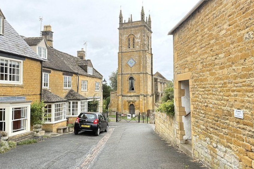 A narrow street with stone buildings, a parked car, and a church with a tall tower. 3