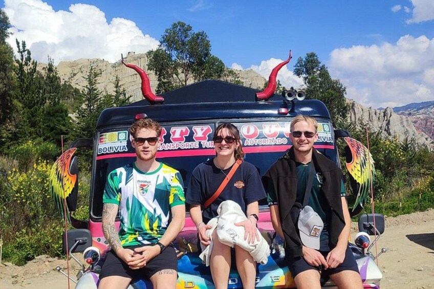 Three people sitting on a colorful bus with a mountainous landscape in the background. 2
