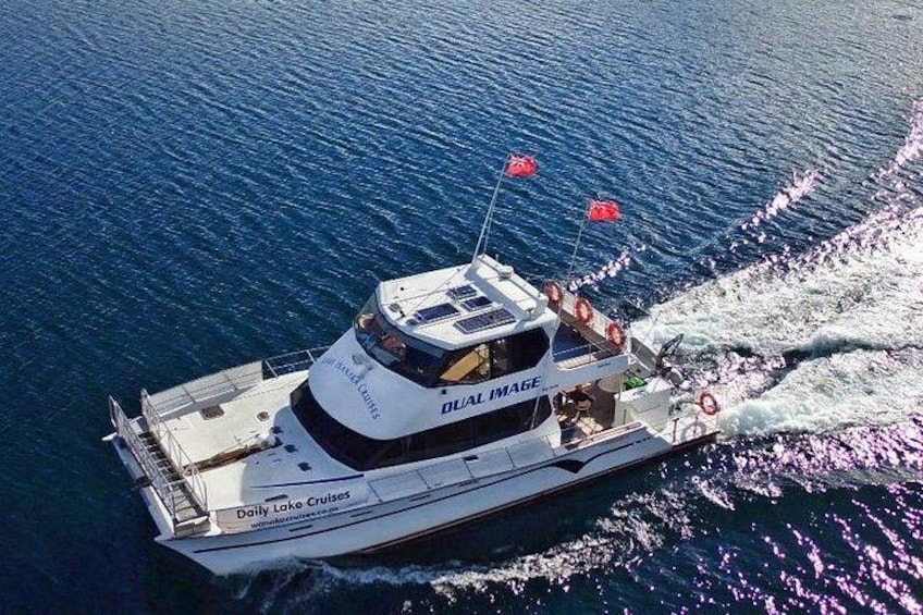 Een witte boot met de tekst 'Daily Lake Cruises' en 'DIAL IMAGE' op de zijkant, die door het water vaart en een kielwalm achterlaat. 5