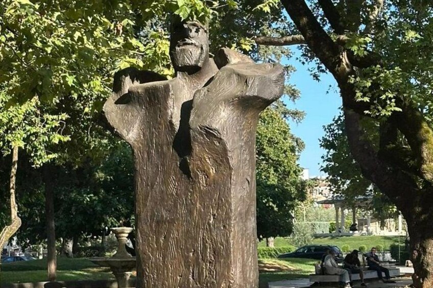 Eine bronzene Statue eines Mannes mit ausgestreckten Armen, eingebettet in einen Park mit Bäumen und einer Bank im Hintergrund. 5