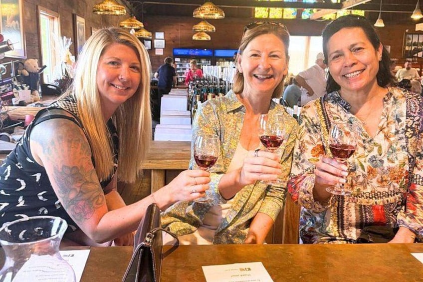 Tres personas sentadas en una mesa con copas de vino, en un restaurante con luces colgantes y muebles de madera. 1