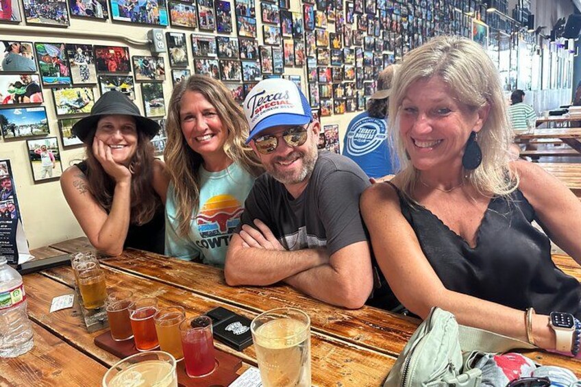A group of people sitting at a wooden table in a bar with various drinks and a wall of photos in the background. 1