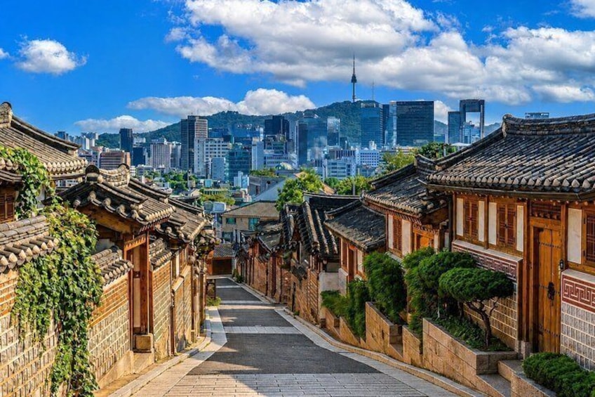 A traditional Korean alley with tiled roofs, leading towards a modern cityscape with skyscrapers and a prominent tower. 1