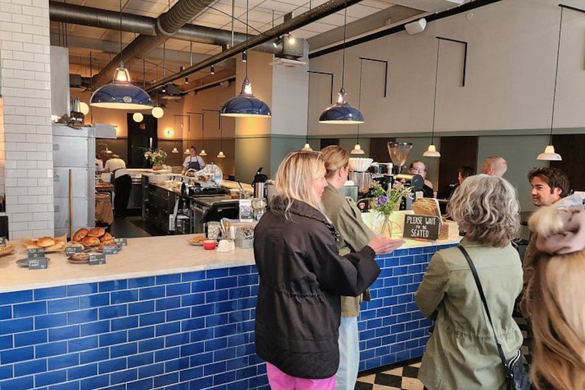 A café interior with a blue tiled counter, hanging pendant lights, and a sign indicating a waitlist. 2