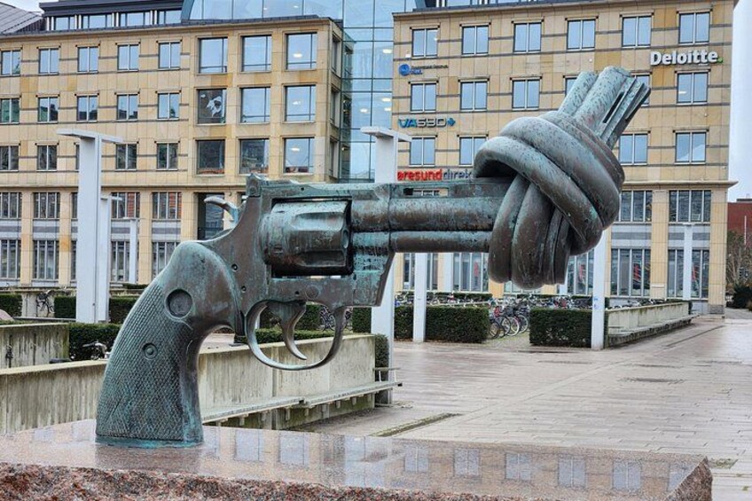 A sculpture of a revolver with a tire on the handle, located in an urban square with buildings in the background. 3