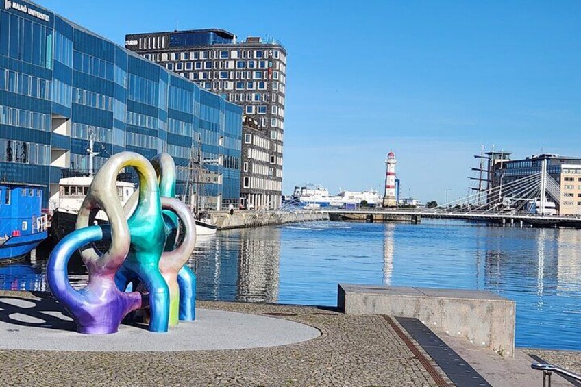 A waterfront with a colorful sculpture, modern buildings, and a lighthouse. 5
