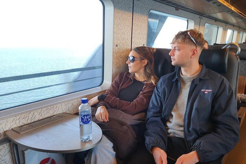 Two people seated by a window on a train, with a water bottle on a table. 2