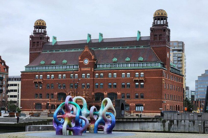 A historic building with a clock tower, a large fountain with colorful sculptures, and a river in the foreground. 2