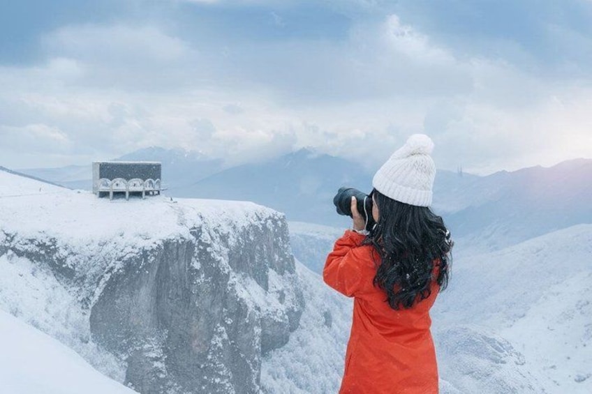 A person in a red jacket taking a photograph of a building on a snowy mountain. 1