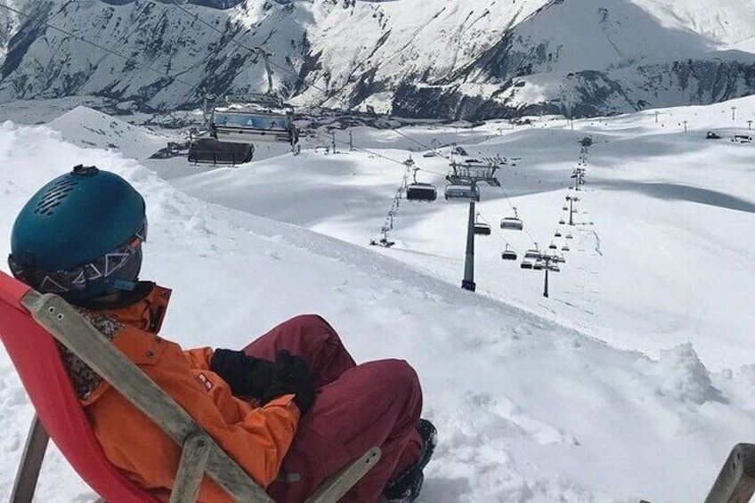 Una persona seduta su una seggiovia in una zona montuosa innevata, con impianti di risalita e pendii ricoperti di neve sullo sfondo. 1