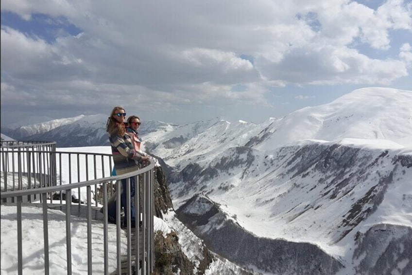 Due persone in piedi su un crinale montuoso innevato, con una ringhiera metallica, che si affaccia su una valle. 3