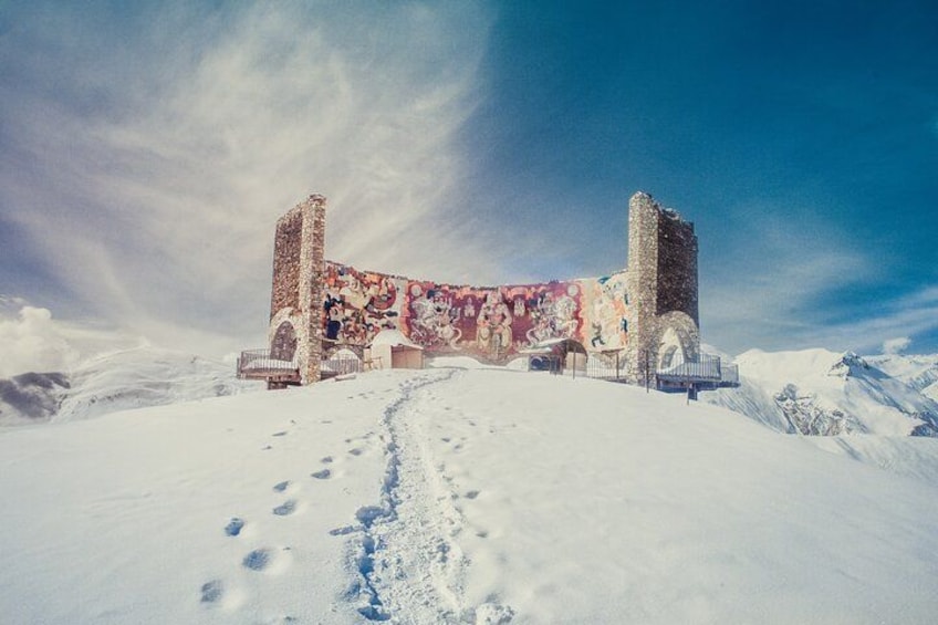 A snow-covered landscape with a historic building featuring colorful murals, a clear sky, and a snow-covered path leading to the structure. 4