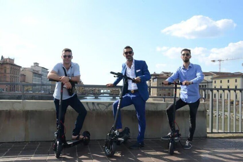Three people on electric scooters on a bridge with buildings in the background. 3