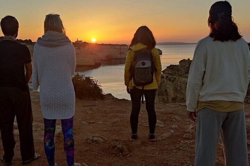 Cuatro personas de pie en un acantilado rocoso contemplando la puesta de sol sobre un cuerpo de agua. 1
