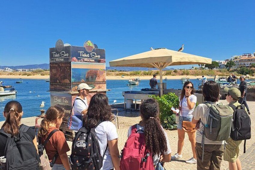 A group of people standing by a ticket shop near a waterfront with boats and a clear blue sky. 5