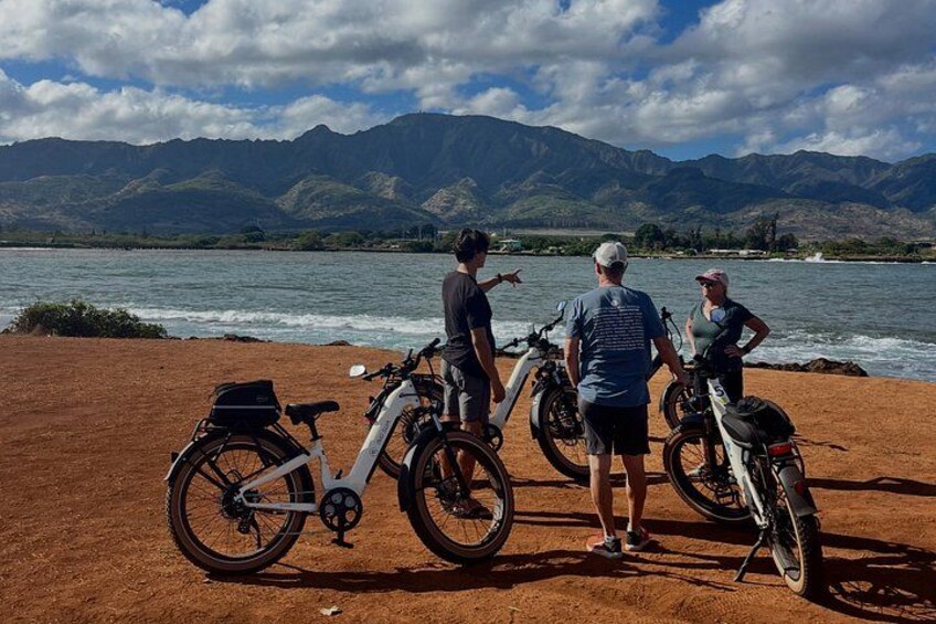 A group of people by a lake with mountains in the background, bicycles, and a dirt path. 1