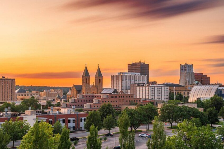 A cityscape at sunset with prominent buildings, including a church with two towers, modern high-rises, and a large glass structure. 2