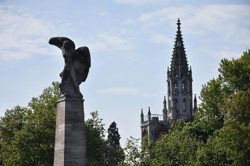 Una estatua de un ángel sobre un pedestal, una iglesia de estilo gótico al fondo, y árboles verdes y frondosos. 4