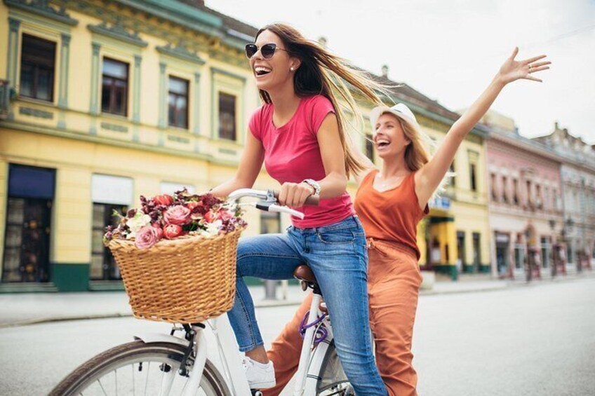 Two people riding a bicycle with a basket of flowers, on a street with buildings. 3