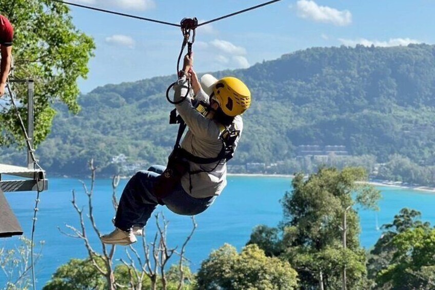 A person zip-lining with a helmet and harness, overlooking a coastal landscape with trees and hills. 1
