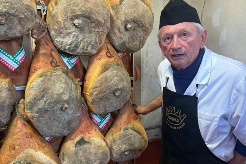 En person står bredvid ett bord med olika sorters lufttorkat kött, personen bär en vit skjorta och ett svart förkläde med en logotyp. 2