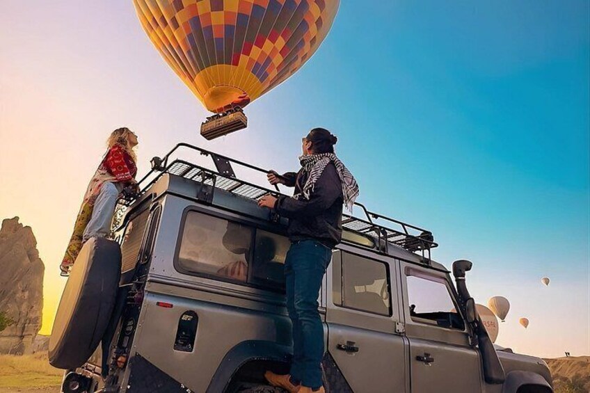 A vehicle with a person standing on the roof, a hot air balloon in the background, and a desert landscape. 1