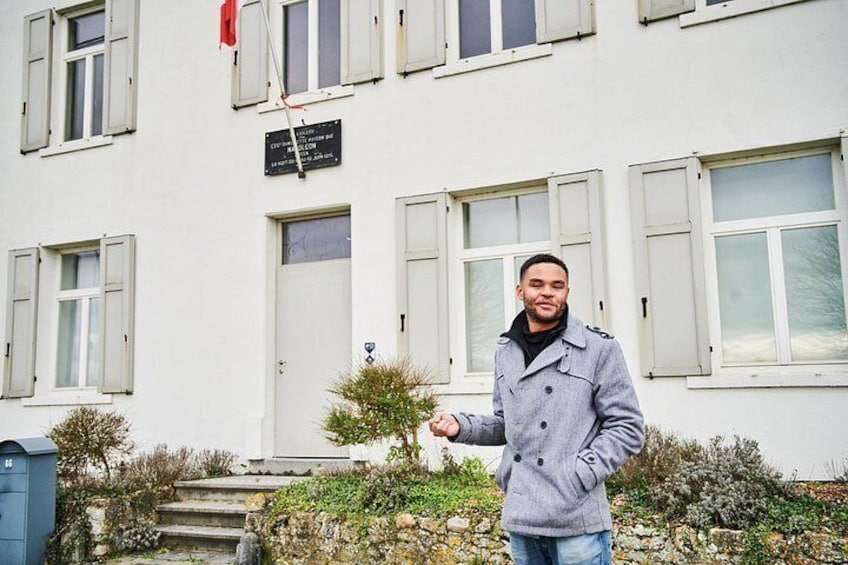 A person standing in front of a white building with shutters, a small tree, and a mailbox. 1