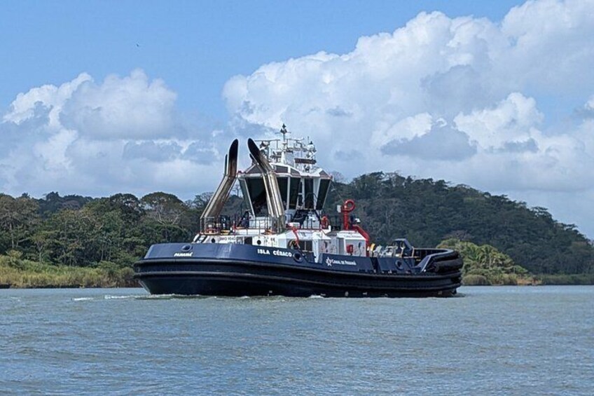 A large black boat with a cabin and equipment on a calm body of water, surrounded by greenery and a clear sky. 2