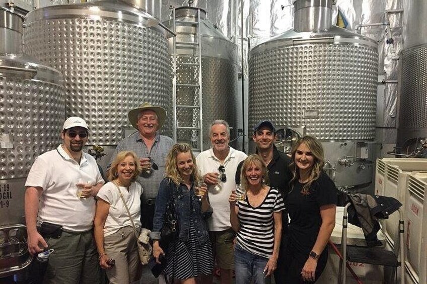A group of people in a winery with large stainless steel fermentation tanks. 5