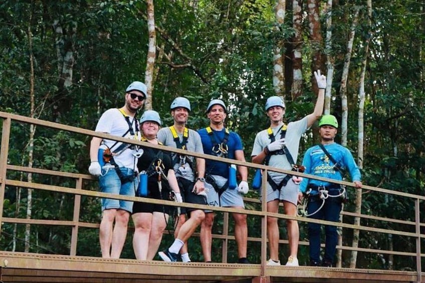 A group of people on a zipline platform, wearing helmets and harnesses, with trees in the background. 4