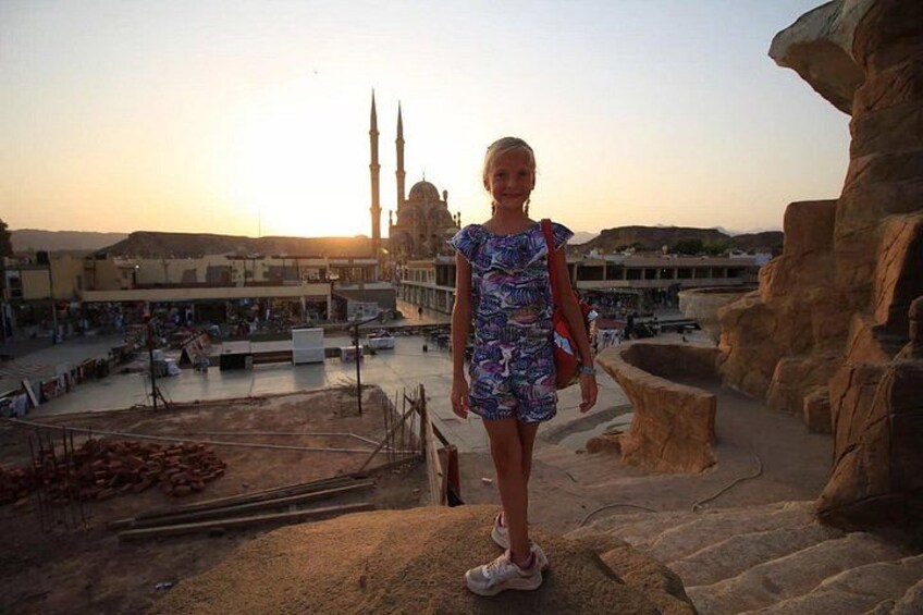 A person standing on a rocky ledge with a mosque and a sunset in the background. 4