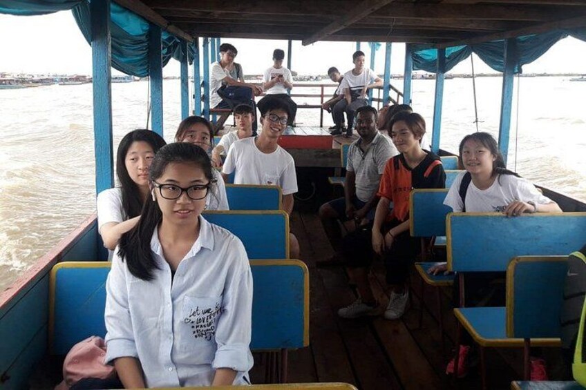 A group of people on a boat with blue and yellow seats, some seated and some standing, with water visible in the background. 3