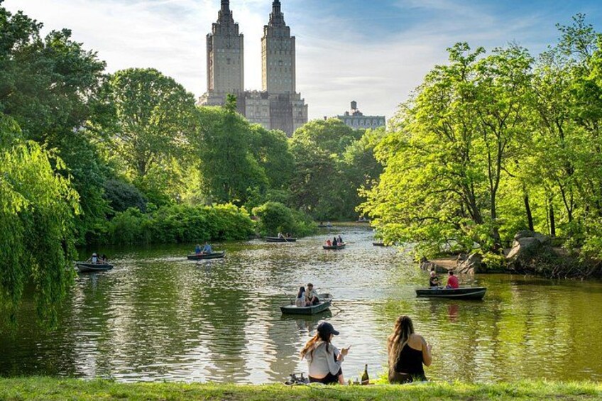 A river with boats, people sitting on the bank, and two prominent buildings in the background. 1
