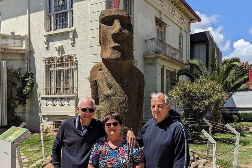 Three people standing in front of a large stone statue with a face, in front of a building with a balcony and a sign. 3