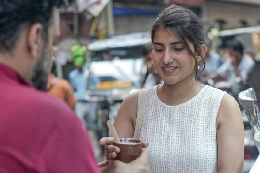 A person in a white sleeveless top holding a small brown container, likely a food item, in an outdoor market setting. 1