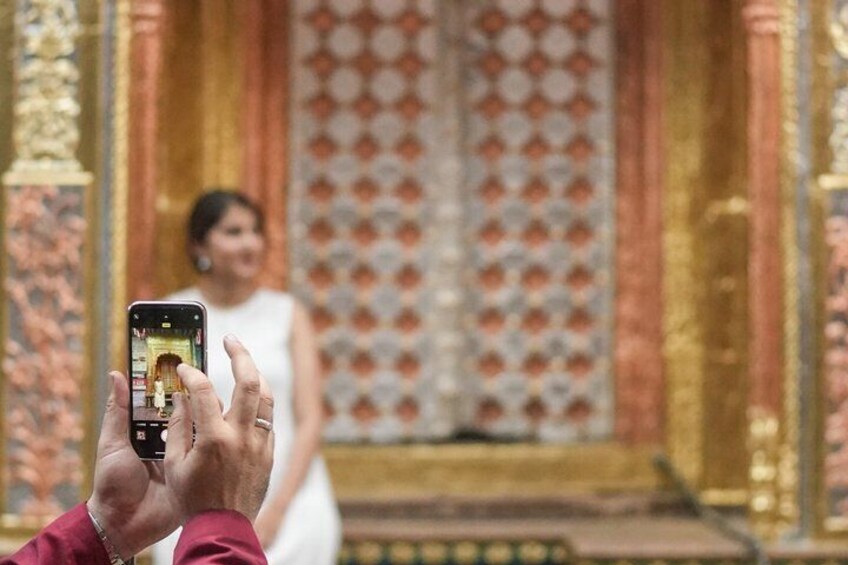 A person taking a photo of a person in a white dress in front of a patterned wall with golden columns. 2
