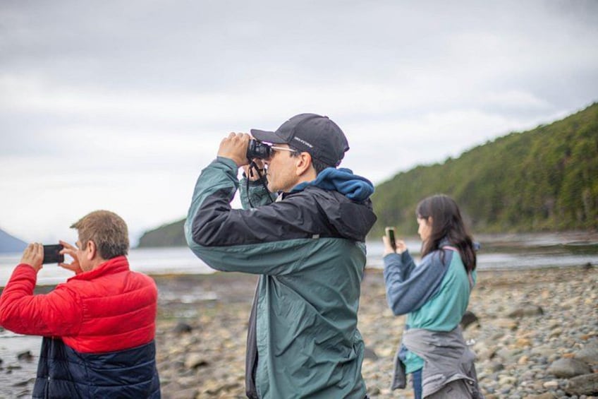 Three people taking photographs by a rocky shore with a body of water and hills in the background. 4