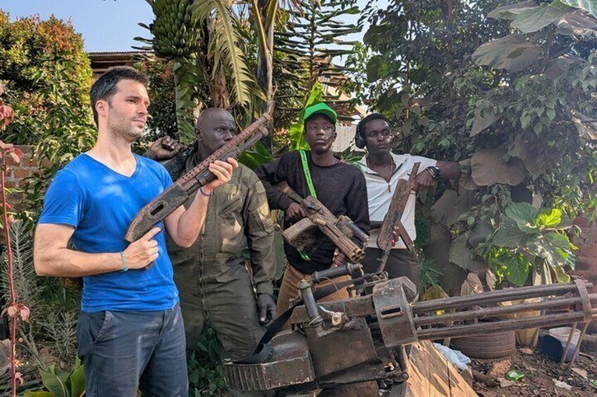 A man holding a large, rusty gun, surrounded by greenery and other people. 3