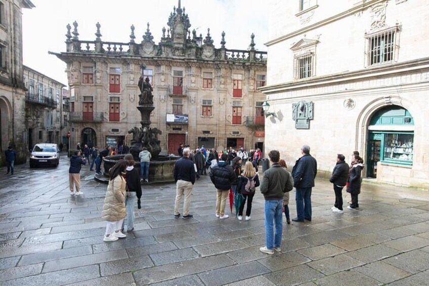Una plaza histórica con una fuente, gente caminando y edificios ornamentados. 1