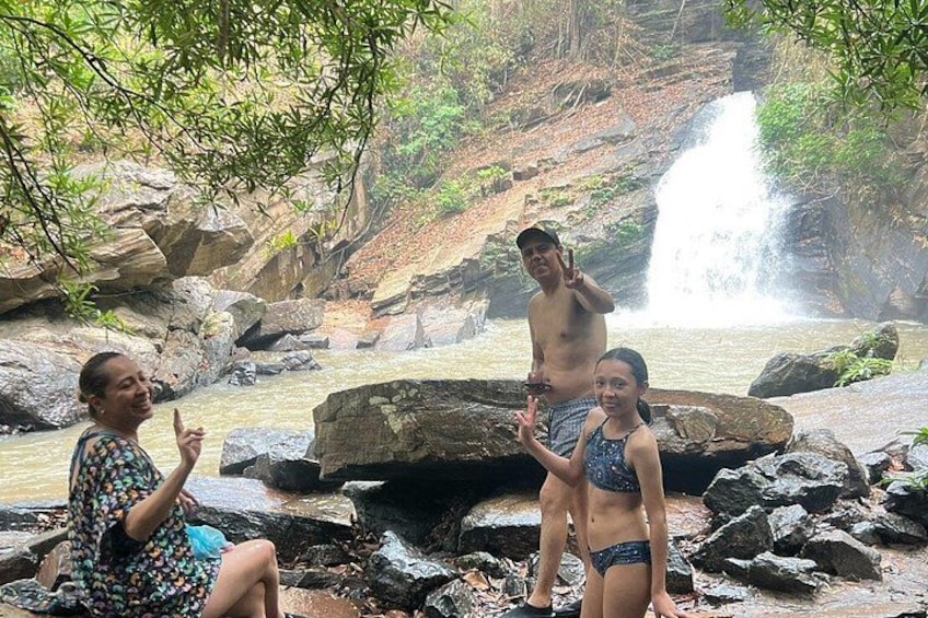 Three people near a waterfall in a forested area with large rocks and a river. 3