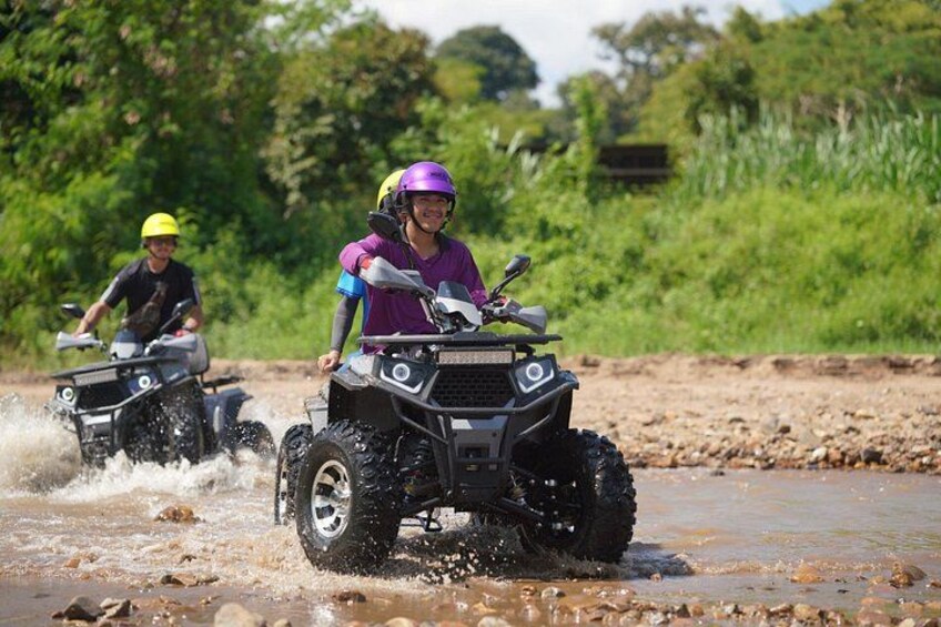 Two people riding ATVs on a muddy path with water splashing around. 1