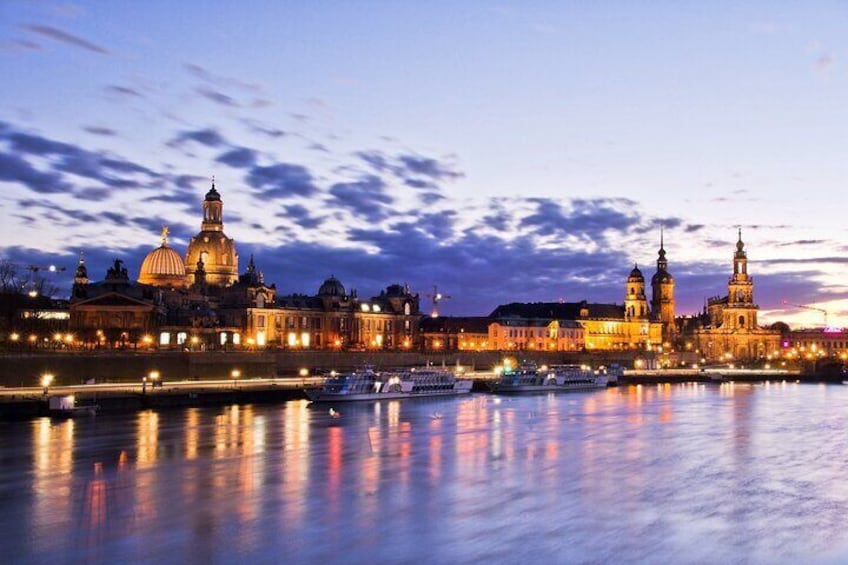 A cityscape at dusk with illuminated buildings and a river reflecting the lights. 1