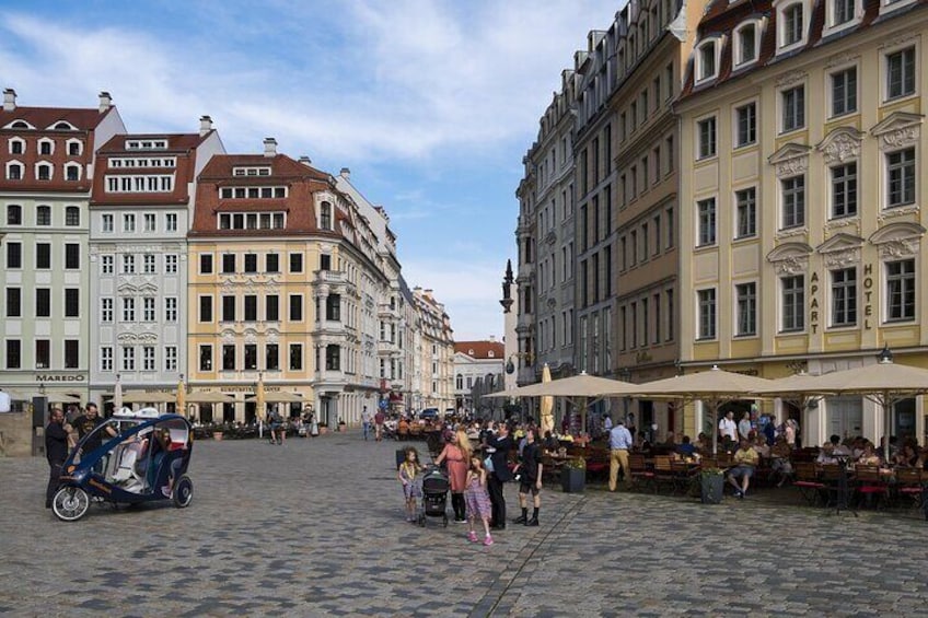 Ett torg med kullerstenar, omgivet av historiska byggnader, med uteserveringar och några få personer. 2