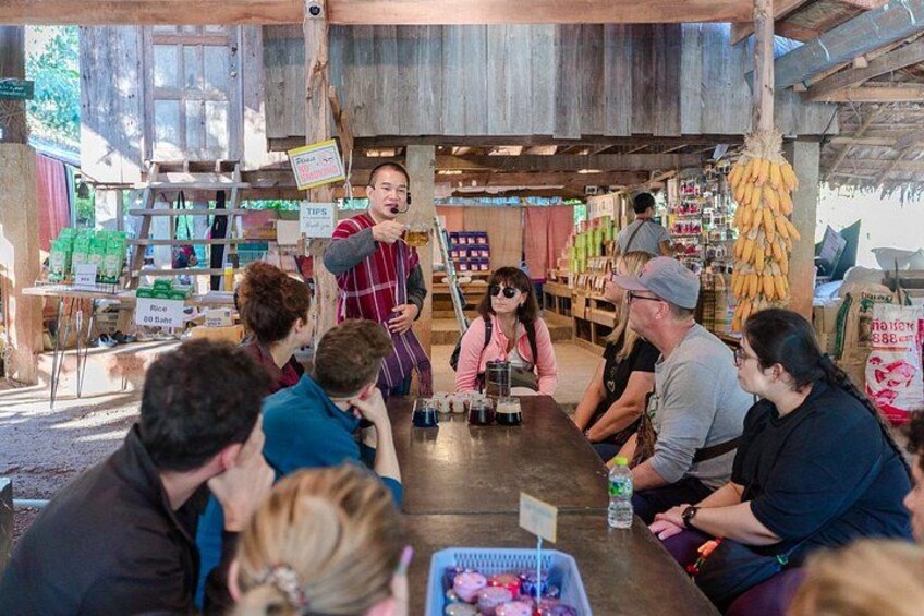 A group of people seated at a wooden table inside a rustic market with various items and signs in the background. 4