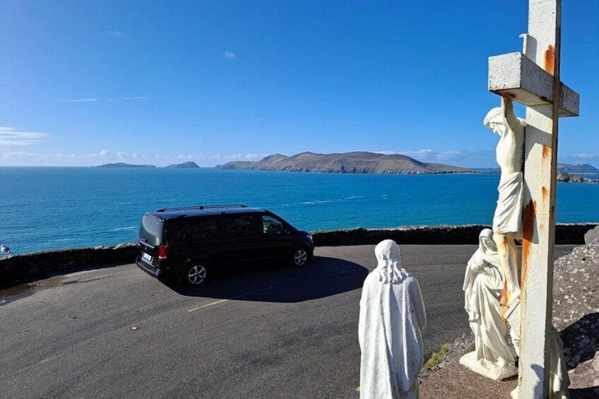 Ein schwarzer Van am Straßenrand, eine weiße Statue einer Person und ein Kruzifix mit einer Figur darauf, mit Blick auf ein Gewässer und ferne Hügel. 5