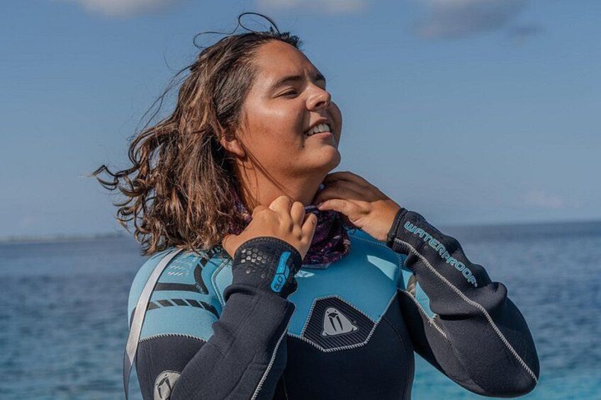 A person wearing a wetsuit with a logo on the sleeve, standing by the ocean. 1