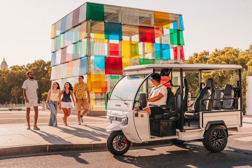 A white electric vehicle with passengers, a colorful building, and a person walking in the background. 4